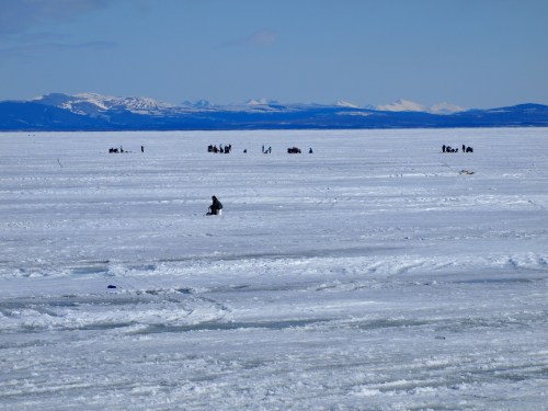 A snowy landscape with people ice fishing on a frozen lake, surrounded by distant mountains under a clear blue sky.