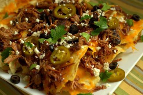 A plate of nachos topped with pulled pork, jalapeños, olives, cheese, and cilantro.