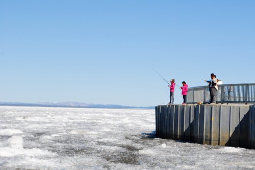 Three people fishing from a pier on a frozen lake, with ice and clear blue skies in the background.