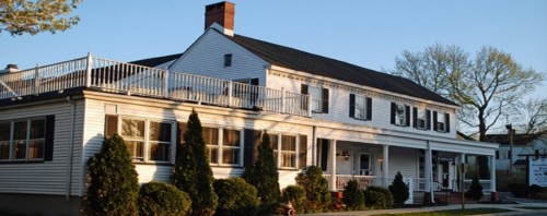 A large white house with a balcony, surrounded by green shrubs and trees, under a clear blue sky.