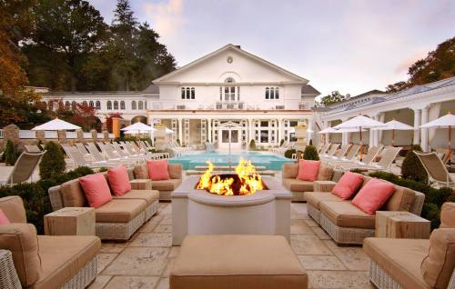 A luxurious outdoor lounge area with a fire pit, surrounded by seating, a pool, and a grand building in the background.