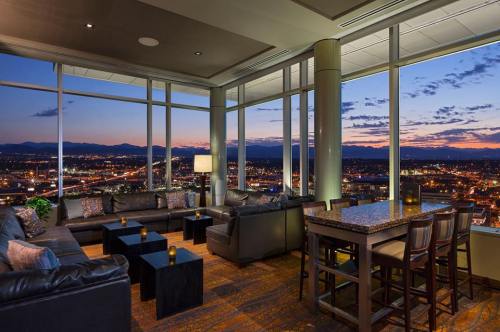 A stylish lounge with large windows showcasing a sunset view over a city skyline, featuring comfortable seating and a bar.