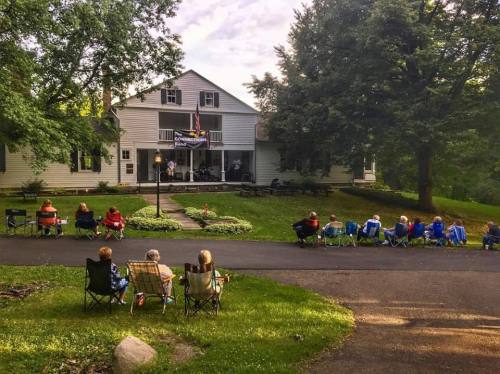 A group of people sitting in lawn chairs facing a house, surrounded by trees and greenery, enjoying an outdoor event.