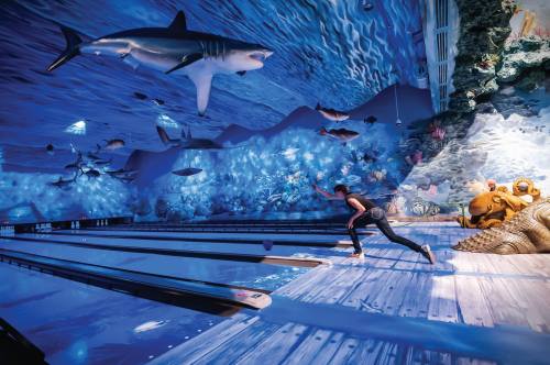 A person bowls in a vibrant, underwater-themed bowling alley with sharks and colorful sea life on the walls.
