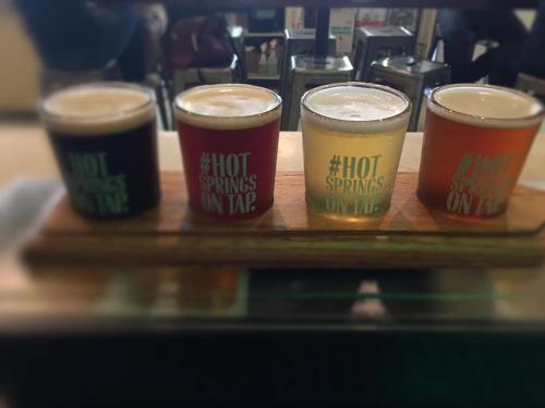 Four glasses of beer in different colors, labeled "#HOT SPRINGS ON TAP," arranged on a wooden tray.