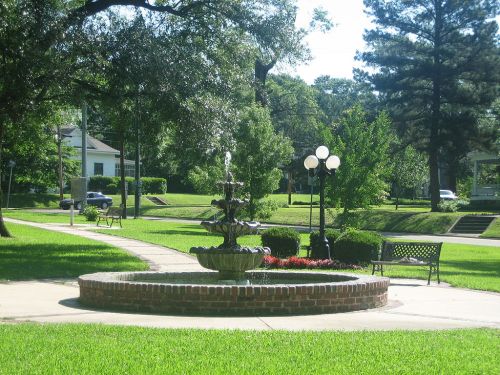 A serene park scene featuring a fountain, benches, and lush greenery under clear skies.