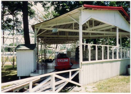 A red roller coaster car named "Cyclone" sits at a wooden loading station surrounded by trees.
