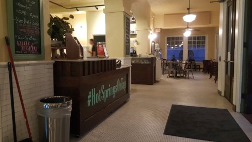 Interior of a restaurant with a wooden counter, seating area, and large windows, featuring a welcoming atmosphere.