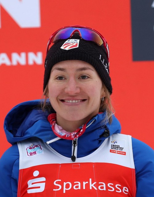 A smiling female athlete in a blue jacket and black hat stands in front of a red backdrop, representing a sports event.