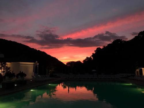 A serene pool at sunset, reflecting vibrant pink and orange hues against a backdrop of dark hills and clouds.