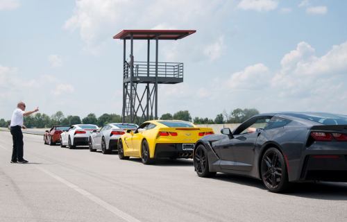 A line of colorful sports cars on a racetrack, with an instructor gesturing from a tower in the background.