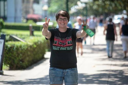 A smiling person in a black t-shirt and shorts holds a water bottle, making a peace sign on a busy sidewalk.