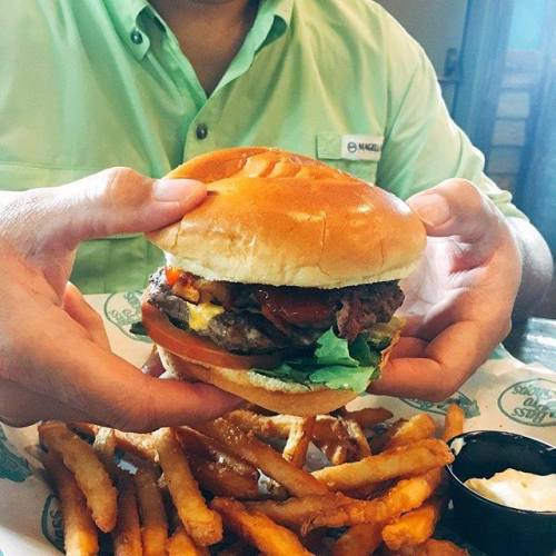A person holding a burger with lettuce, cheese, and bacon, accompanied by a side of fries and dipping sauce.