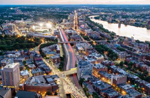 Aerial view of a cityscape at dusk, featuring buildings, a river, and light trails from moving vehicles.