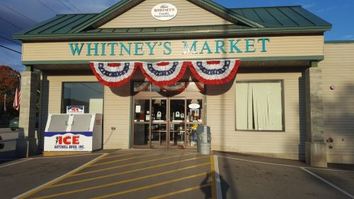 Exterior of Whitney's Market, featuring patriotic bunting and a sign, with an ice vendor in front.