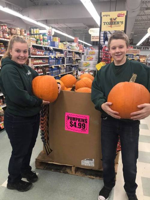 Two young people hold large pumpkins in a grocery store aisle, with a sign reading "PUMPKINS $4.99" in the background.