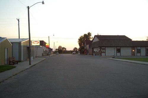 Quiet street scene at dusk, featuring buildings on either side and a clear sky.