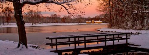 A serene winter scene with a snowy landscape, a frozen lake, and a colorful sunset reflecting on the water.