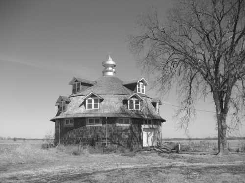 A unique, round barn with multiple windows stands beside a bare tree in a rural landscape, captured in black and white.