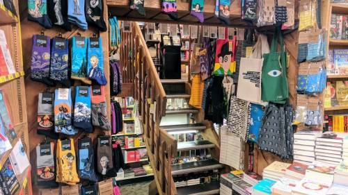A cozy bookstore interior featuring colorful socks, bags, and books displayed on wooden shelves and a staircase.