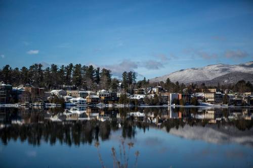 A serene winter landscape featuring a snowy lakeside town reflected in calm waters under a clear blue sky.