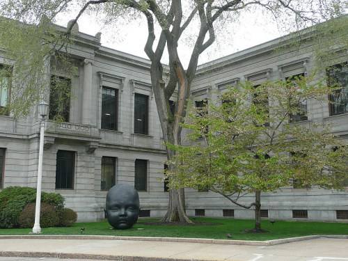 A large black sculpture of a child's face stands in front of a historic stone building, surrounded by trees.