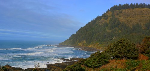 Coastal landscape featuring waves crashing on rocks, lush greenery, and a forested hillside under a blue sky.