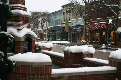 Snow-covered street with brick benches, festive decorations, and shops in a quaint town during winter.