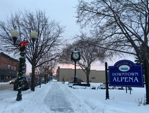 Snowy downtown Alpena with a welcome sign, decorated trees, and a clock, under a pastel sky at dusk.