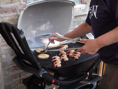 A person grilling skewered shrimp and vegetables on a barbecue grill outdoors.