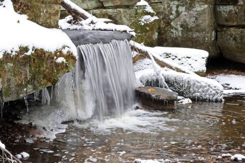 A small waterfall flows over rocks, surrounded by snow and ice in a serene, winter landscape.
