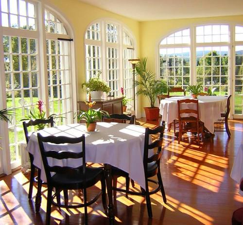 Bright sunlit room with large windows, featuring tables with white tablecloths and potted plants.