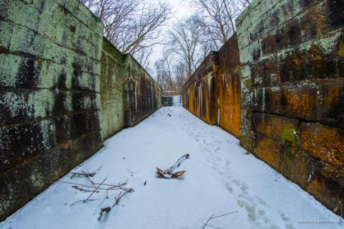 A snow-covered path between weathered stone walls, surrounded by bare trees in a wintry landscape.