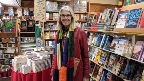 A smiling woman with long hair stands in a cozy bookstore surrounded by shelves of books and a table of stacked novels.