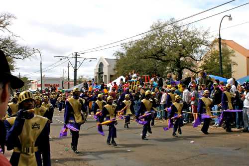 A lively parade scene with performers in gold and purple costumes dancing, surrounded by a cheering crowd.