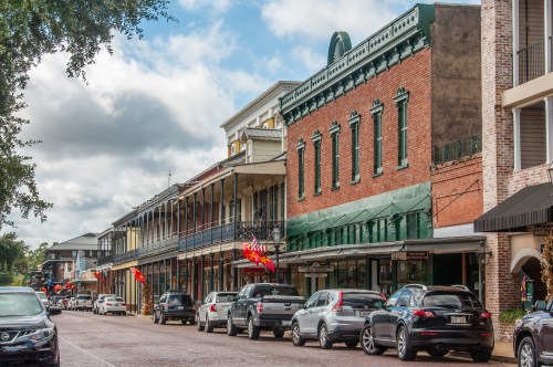 Historic buildings line a street with parked cars, under a cloudy sky in a charming town.