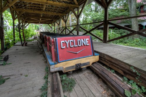 An abandoned roller coaster car labeled "Cyclone" sits on a wooden track under a rustic shelter, surrounded by greenery.