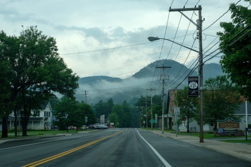 A quiet road lined with trees, leading to misty mountains under a cloudy sky. Power lines run alongside the street.