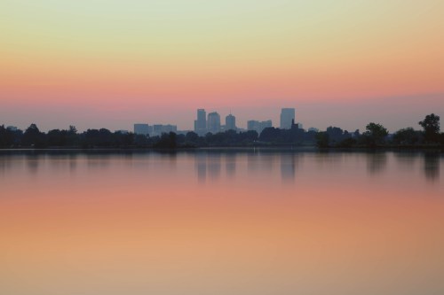 A serene lake reflects a pastel sunset with a city skyline in the background.