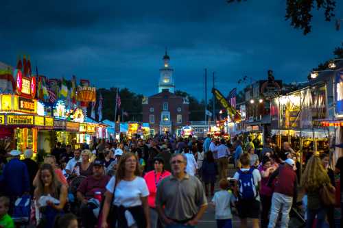 A bustling fairground at dusk, filled with people, colorful stalls, and a clock tower in the background.