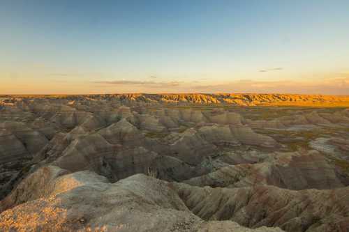 A panoramic view of layered rock formations in a vast, arid landscape during sunset, with soft golden light illuminating the scene.