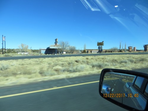 A view of a highway with dry grass, roadside signs, and distant buildings under a clear blue sky.