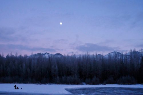 A serene winter scene with a person ice fishing on a frozen lake, surrounded by mountains and a twilight sky.
