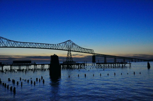 Silhouette of a long bridge over calm water at dusk, with a gradient sky transitioning from blue to orange.