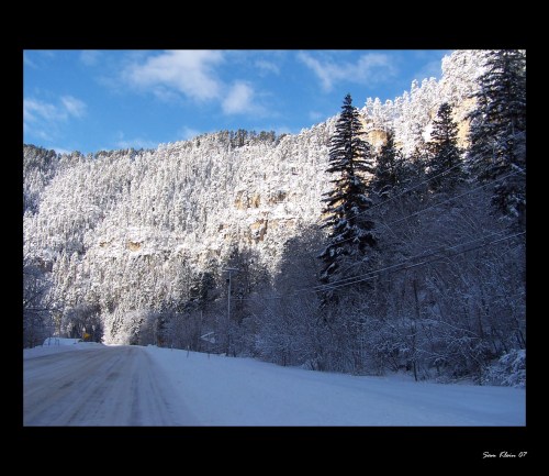 Snow-covered landscape with trees and mountains under a blue sky, along a quiet, snowy road.