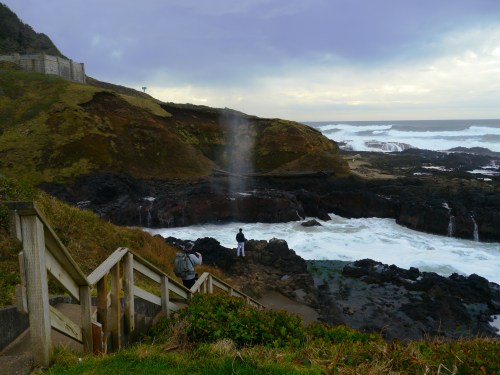 A coastal scene with waves crashing on rocks, a person standing near a blowhole, and a staircase leading down to the shore.
