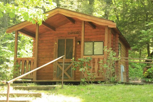 A small wooden cabin surrounded by trees, featuring a porch and steps leading up to the entrance.