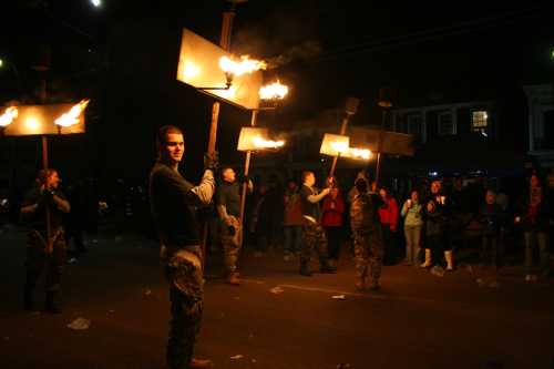 A group of people holding torches march down a street at night, with onlookers watching from the sidelines.