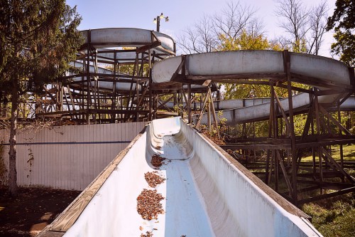 Abandoned water slide with overgrown foliage and debris, set against a clear blue sky.