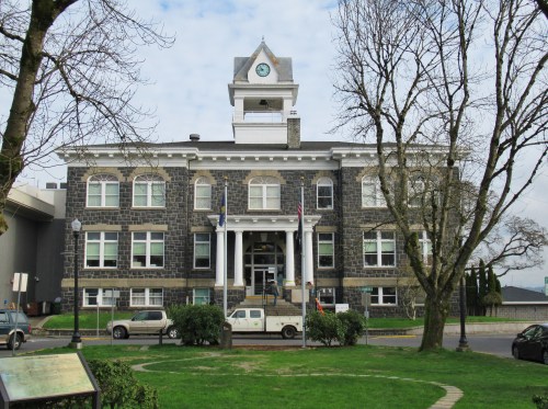 Historic stone building with a clock tower, surrounded by trees and a grassy area, under a cloudy sky.